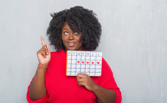 Young african american woman over grey grunge wall holding menstruation calendar surprised with an idea or question pointing finger with happy face, number one