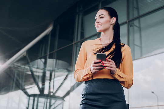 Positive expectation. Beautiful lady is standing near the entrance and waiting with smartphone. Copy space in left side