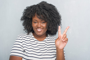 Young african american plus size woman over grey grunge wall smiling with happy face winking at the camera doing victory sign. Number two.