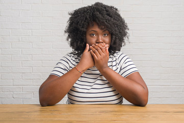 Young african american woman sitting on the table at home shocked covering mouth with hands for...