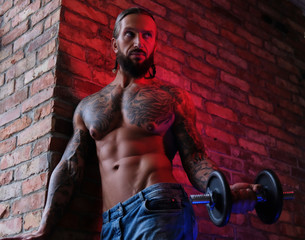 Brutal bearded tattooed male doing exercise with a dumbbell in a room with loft interior.