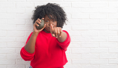 Young african american woman over white brick wall eating avocado pointing with finger to the camera and to you, hand sign, positive and confident gesture from the front