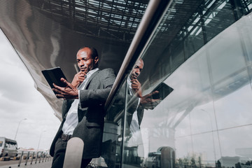 Lets think. Low angle portrait of young male leaning on glass fence at the airport. Man is holding tablet with pensive look