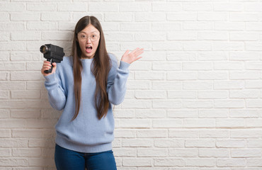 Young Chinese woman filming holding video camera very happy and excited, winner expression celebrating victory screaming with big smile and raised hands