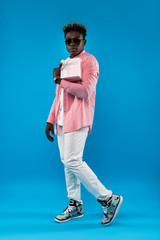 Best wishes. Full length portrait of young man in sunglasses holding pink gift box with ribbon. Isolated on blue background