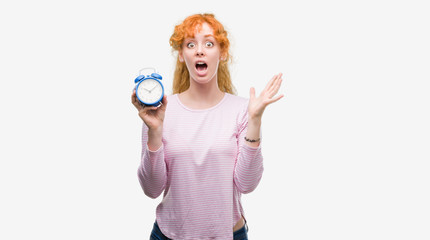 Young redhead woman holding alarm clock very happy and excited, winner expression celebrating victory screaming with big smile and raised hands