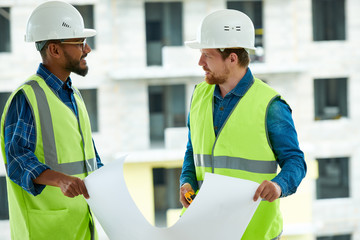 Serious pensive young multiethnic engineers in white hardhats and safety work vests discussing building project while viewing blueprint at construction site of apartment building