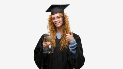 Young redhead student woman wearing graduated uniform holding hourglass happy with big smile doing ok sign, thumb up with fingers, excellent sign