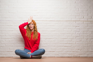 Young redhead woman sitting over brick wall surprised with hand on head for mistake, remember error. Forgot, bad memory concept.