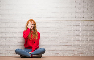 Young redhead woman sitting over brick wall covering one eye with hand with confident smile on face and surprise emotion.