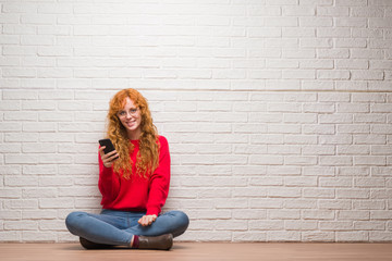 Young redhead woman sitting over brick wall talking on the phone with a happy face standing and smiling with a confident smile showing teeth