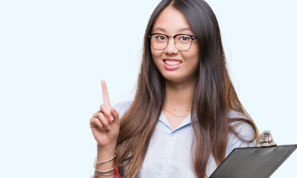 Young asian business woman holding clipboard over isolated background surprised with an idea or question pointing finger with happy face, number one
