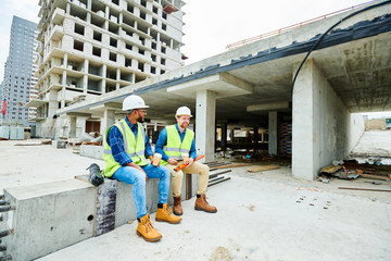Positive handsome young builders in safety vests and hardhats sitting on cement beam and talking...