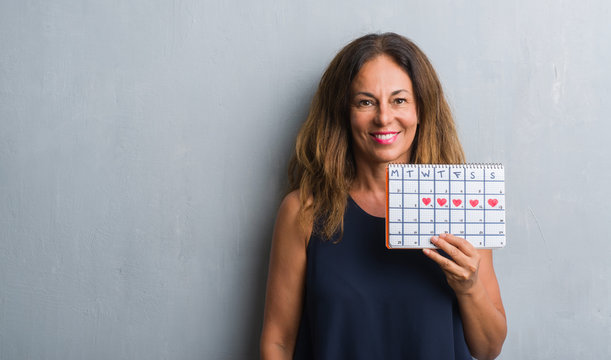 Middle Age Hispanic Woman Standing Over Grey Grunge Wall Holding Period Calendar With A Happy Face Standing And Smiling With A Confident Smile Showing Teeth