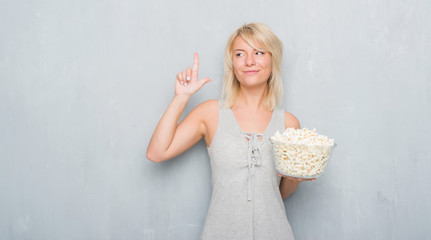 Adult caucasian woman over grunge grey wall eating pop corn surprised with an idea or question pointing finger with happy face, number one