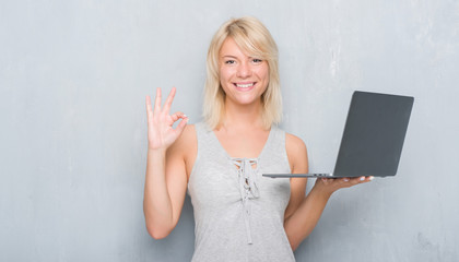 Adult caucasian woman over grunge grey wall using computer laptop doing ok sign with fingers, excellent symbol