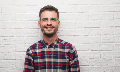 Young adult man standing over white brick wall with a happy face standing and smiling with a confident smile showing teeth