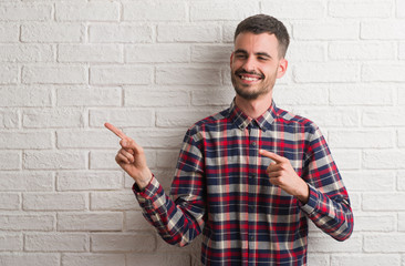 Young adult man standing over white brick wall smiling and looking at the camera pointing with two hands and fingers to the side.