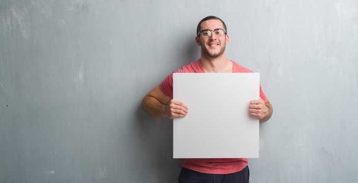 Young caucasian man over grey grunge wall holding a blank banner with a happy face standing and smiling with a confident smile showing teeth
