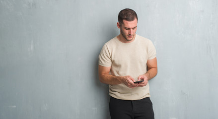 Young caucasian man over grey grunge wall texting a message using smartphone with a confident...