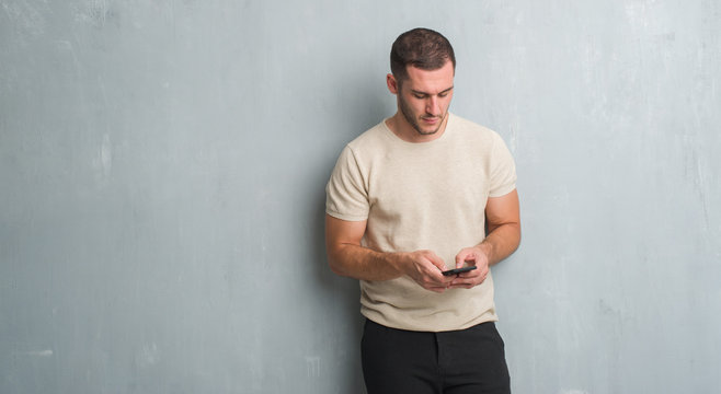 Young caucasian man over grey grunge wall texting a message using smartphone with a confident expression on smart face thinking serious