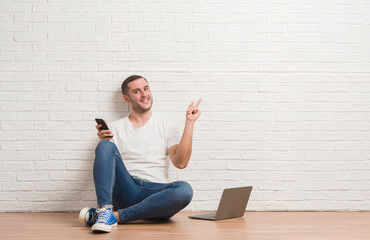 Young caucasian man sitting over white brick wall using computer laptop and smartphone very happy pointing with hand and finger to the side