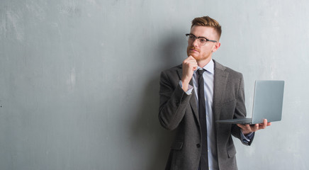Young redhead elegant business man over grey grunge wall using laptop serious face thinking about...