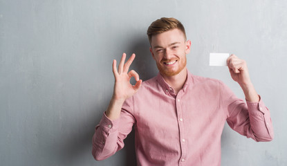 Young redhead man over grey grunge wall holding blank visit card doing ok sign with fingers, excellent symbol