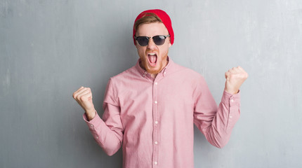 Young redhead man over grey grunge wall wearing wool cap and sunglasses annoyed and frustrated shouting with anger, crazy and yelling with raised hand, anger concept