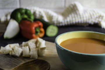 Gazpacho, tomato soup, on top of a wooden board and wooden spoon, with pieces of bread and in the background peppers, onion and cucumber.