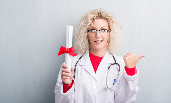 Young blonde doctor woman over grunge grey wall holding degree pointing and showing with thumb up to the side with happy face smiling