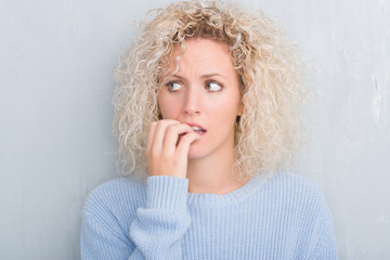 Fototapeta na wymiar Young blonde woman with curly hair over grunge grey background looking stressed and nervous with hands on mouth biting nails. Anxiety problem.