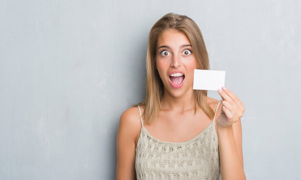 Beautiful young woman over grunge grey wall holding blank visit card scared in shock with a surprise face, afraid and excited with fear expression