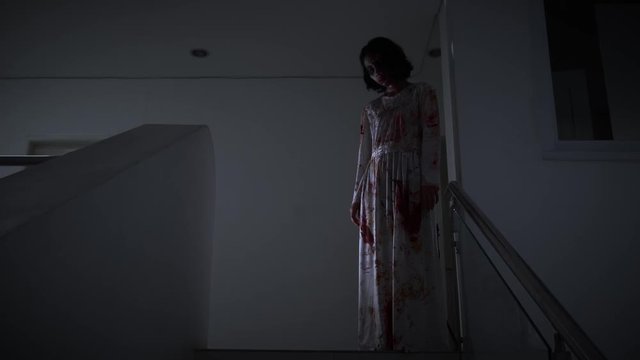 Creepy ghost girl with bloody clothes standing on the stairs at dark night in home. Halloween concept. Shot in 4k resolution