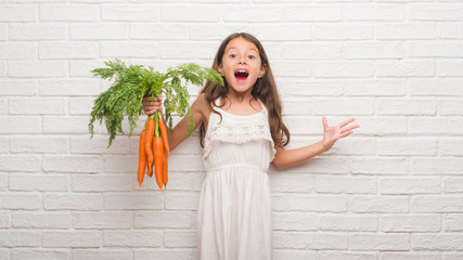 Young hispanic kid over white brick wall holding fresh carrots very happy and excited, winner expression celebrating victory screaming with big smile and raised hands