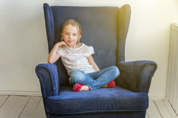 Sweet little girl in jeans and white T-shirt at home sitting on modern cozy blue chair relaxing in white living room. Childhood, schoolchildren, youth, relax concept