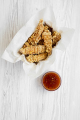 Chicken strips in paper box with sweet chili sauce on a white wooden table, overhead. Flat lay, top view, from above.