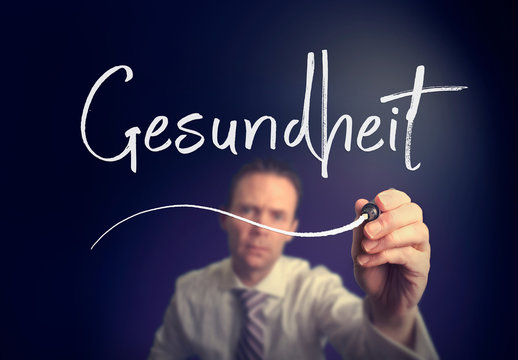 A businessman writing a Health "Gesundheit" concept in German with a white pen on a clear screen.