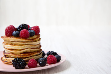 Pancakes with berries on a pink plate over white wooden background, side view. Copy space.