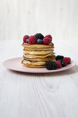 Pancakes with berries on a pink plate over white wooden background, side view.