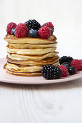 Pancakes with berries on a pink plate over white wooden background, side view. Close-up.