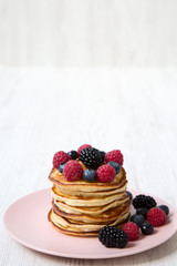 Pancakes with berries on a pink plate over white wooden surface, side view. Space for text.