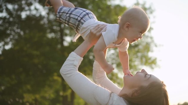 American mother is having fun with cute son in green park in summer, close view of young woman holds infant in hands, relax, standing on background of tree in open air. Concept: two person, freedom