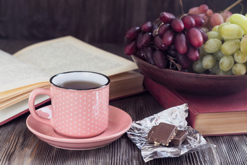 A cup of tea in a pink cup next to an open book and grapes in a plate on a pile of books