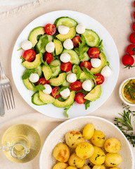 Fresh salad with tomatoes cucumbers arugula mozzarella and avocado. Oil with spices, baked potatoes, top view, vertical