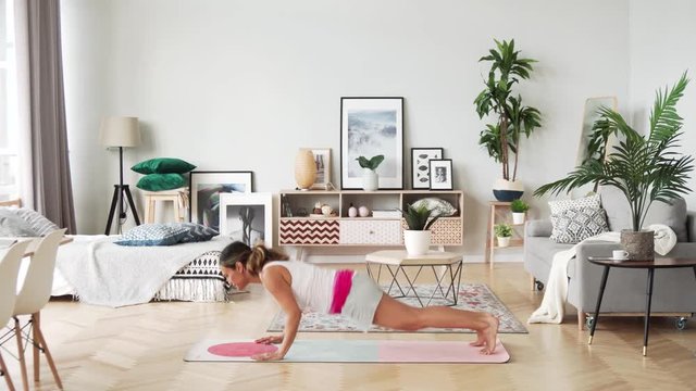 Beautiful slim woman in thirties with fair complexion does morning strenth and aerobic burpee exercises in modern airy room with fair sophisticated interior in Scandinavian style