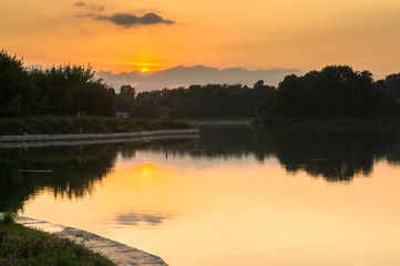 Golden sunset on a pond. Mysterious moment when the orange sun says goodbye to nature. Its last reflection trembles on the water. Evening twilight becomes darker more and more.