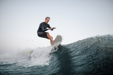 Active man wakesurfing on the board down the blue wave against the background of clear sky