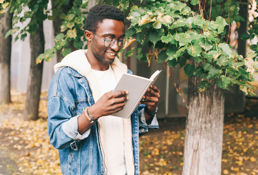 Happy young smiling african man reading book in autumn city park