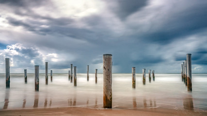 Naklejka na ściany i meble Long shutter photo of wooden on sandy beach with white clouds and waves of the North Sea